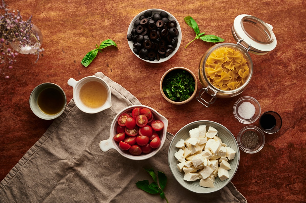 the ingredients for caprese pasta salad on a table: mozzarella, black olives, pasta, olive oil, balsamic vinegar, cherry tomatoes.  