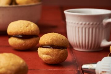 baci di dama on top of a red brick table beside a cup of coffee and a dripping chocolate from a piping tip.