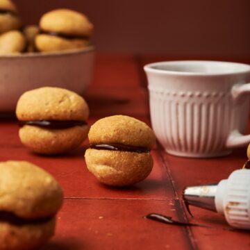 baci di dama on top of a red brick table beside a cup of coffee and a dripping chocolate from a piping tip.