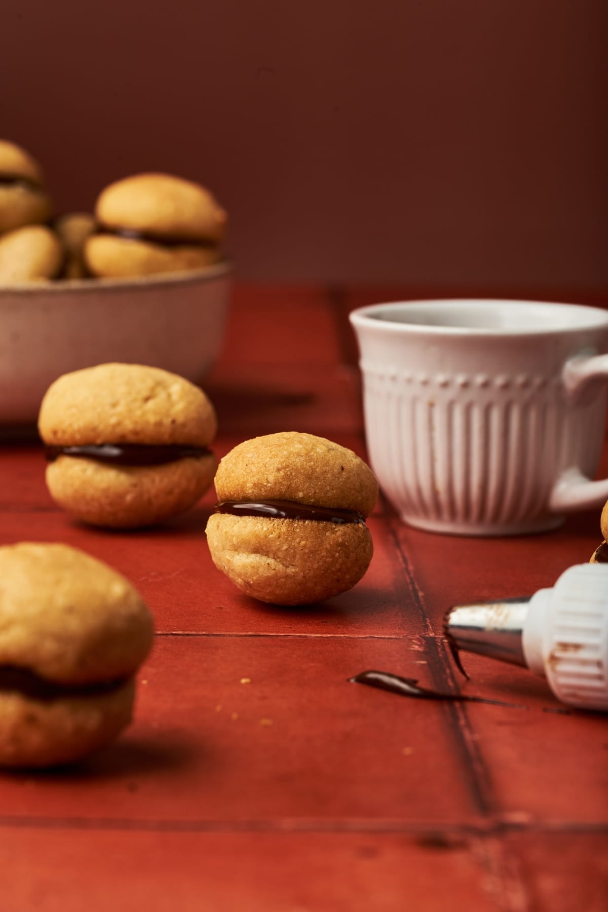 baci di dama on top of a red brick table beside a cup of coffee and a dripping chocolate from a piping tip.