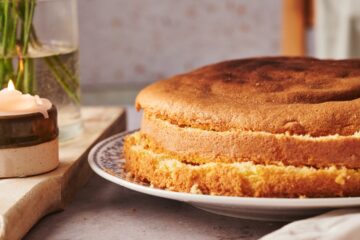italian sponge cake cut into three layers on a plate on top of a marble counter top with a candle and a vase of flowers on top of a wooden chopping board.