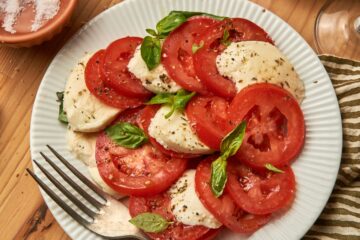 a dish of caprese salad on a white plate with a fork on top of a kitchen towel. next to a larger plate of caprese salad, bowl of salt, some wine glass.