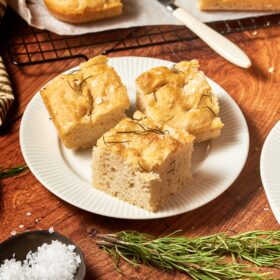 focaccia bread on a white plate with a cooling rack with more bread, some sprigs of rosemary and salt around the plate.