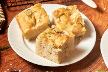 focaccia bread on a white plate with a cooling rack with more bread, some sprigs of rosemary and salt around the plate.