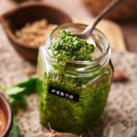 a clear jar of pesto sauce and a spoonful of pesto. around the jar are some garlic cloves, wooden bowls, pine nuts and fresh basil.