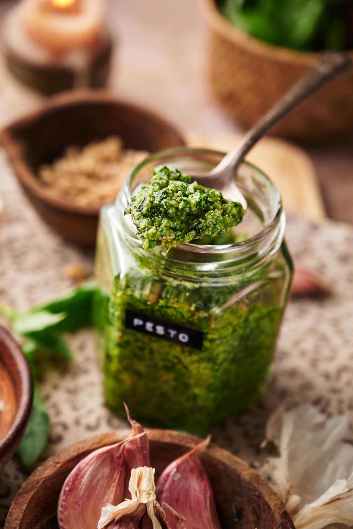 a clear jar of basil pesto sauce and a spoonful of pesto. around the jar are some garlic cloves, wooden bowls, pine nuts and fresh basil.