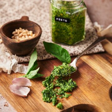 clear jar of basil pesto with a black label with white text labeled pesto. beside a pestle with pine nuts inside. some fresh basil leaves and garlic peel scattered around. and a spoon of pesto.