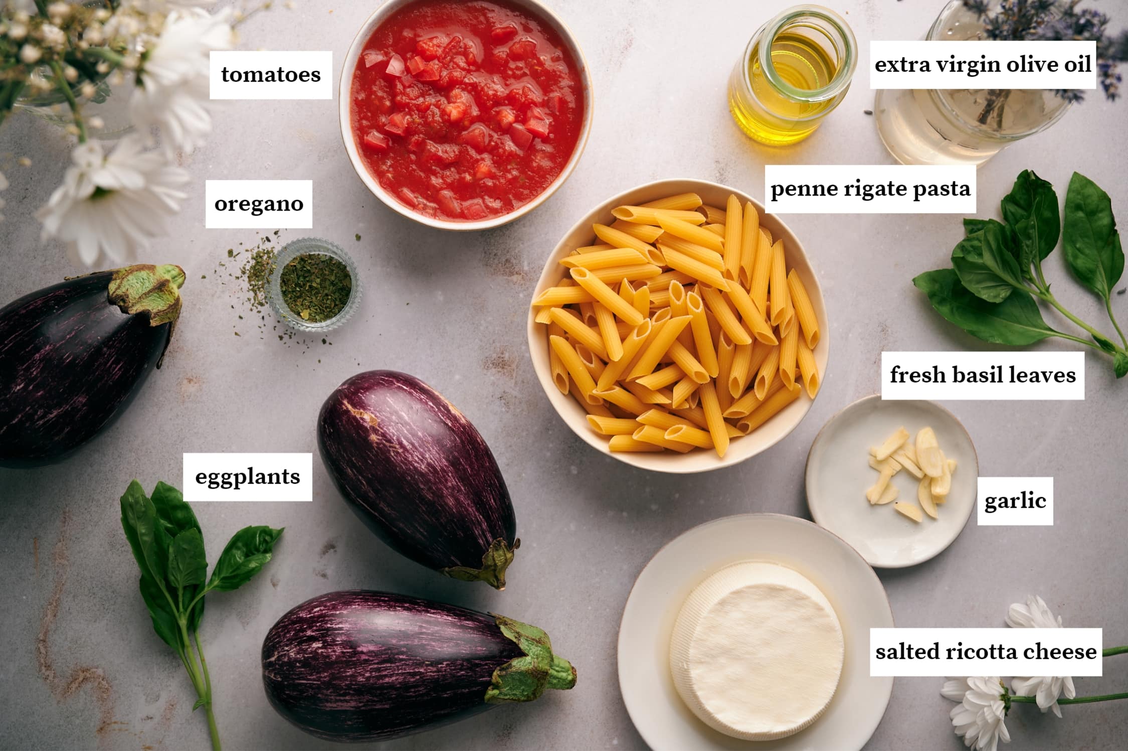 ingredients of pasta alla norma: bowl of tomatoes, bottle of olive oil, fresh basil, penne pasta in a white bowl, oregano, eggplants, sliced garlic on a small plate, salted ricotta cheese on a white plate.