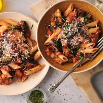 pasta alla norma on an orange dish on top of a wooden chopping board, next to a plate of pasta alla norma in a white plate.