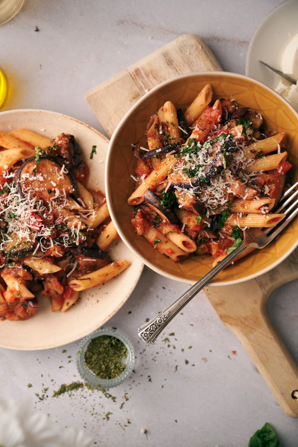 pasta alla norma on an orange dish on top of a wooden chopping board, next to a plate of pasta alla norma in a white plate.